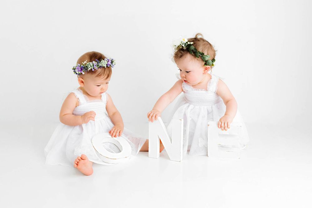 Two baby girls in white dresses and floral crowns playing with ONE letters during a first birthday photo session.