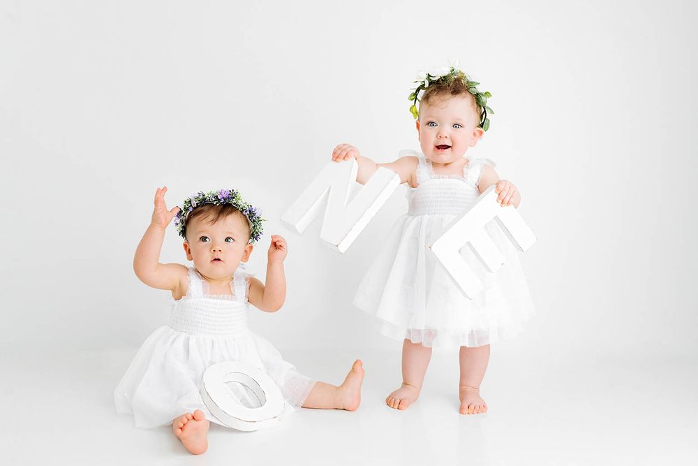Two baby girls in white dresses and floral crowns holding white clothing items against a white background.