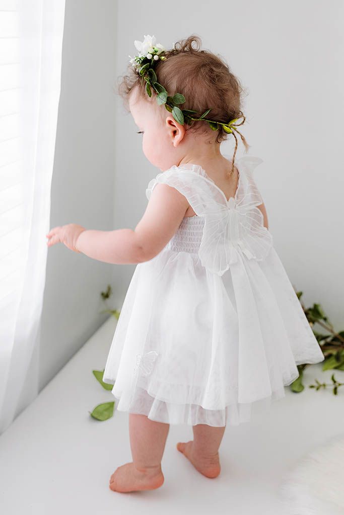 Toddler girl in white tulle butterfly dress and floral crown stands by curtain, viewed from behind.