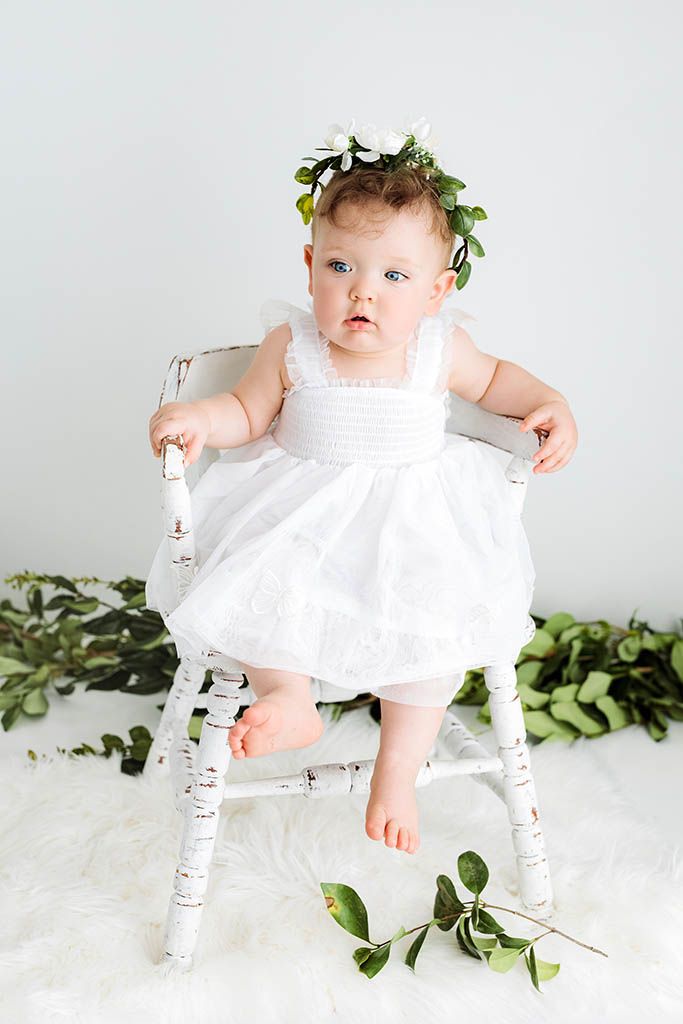 Baby girl in white dress and floral crown sits on white chair surrounded by green leaves.