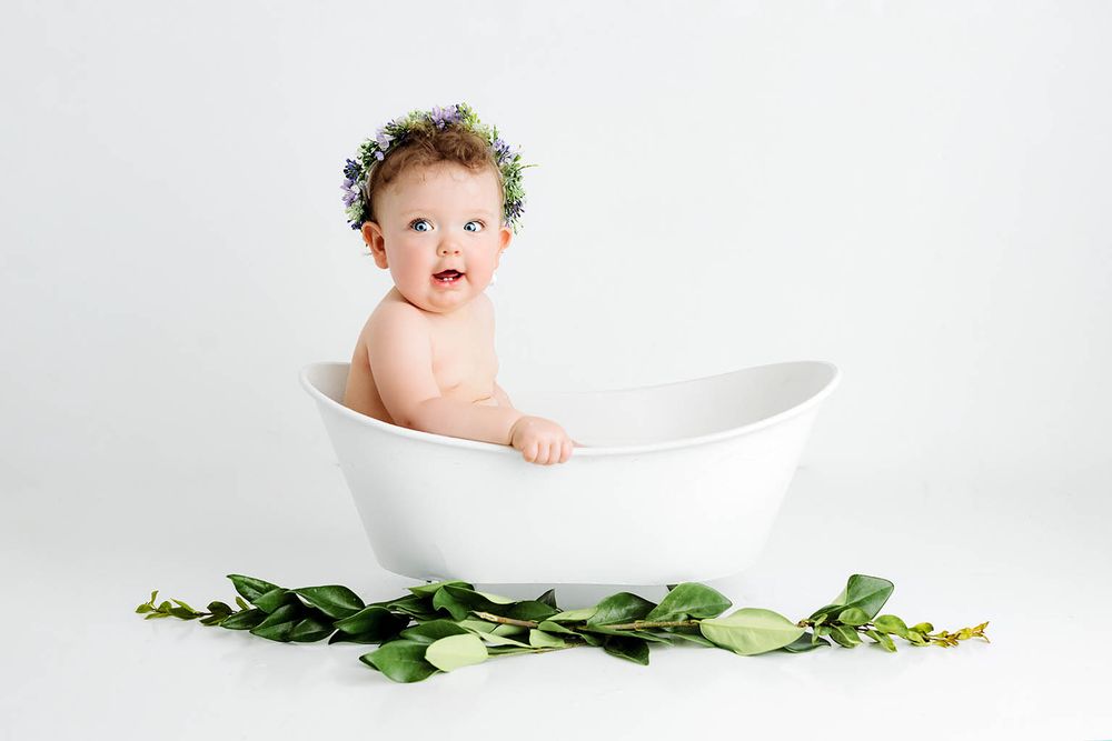 Adorable baby wearing a floral crown sitting in a white bathtub surrounded by green leaves.