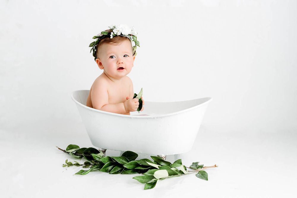 Adorable baby sits in white bathtub with green leaf crown, surrounded by botanical greenery.