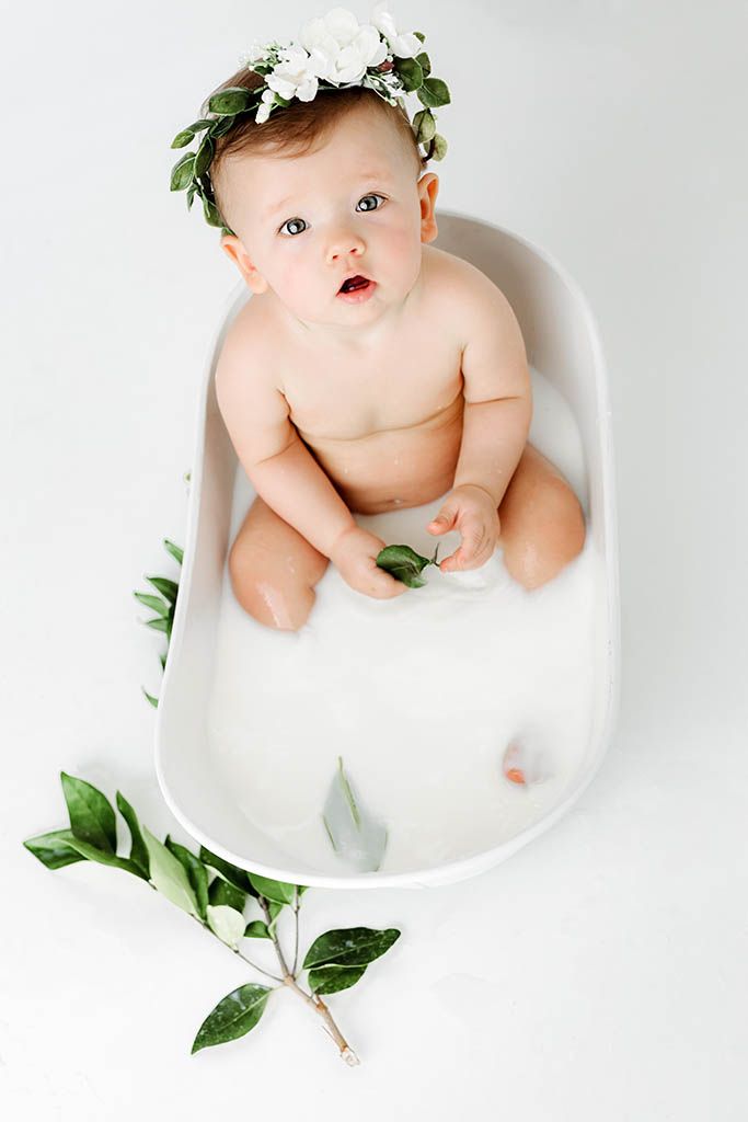 Baby with white floral crown sits in milk bath tub, looking up, with green branches nearby.