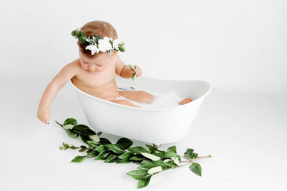 Sweet baby wearing white flower crown plays in milk bath tub with green leafy branches.
