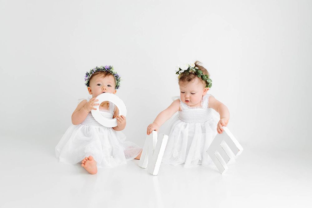 Two baby girls in white dresses and floral crowns playing with large white alphabet letters.