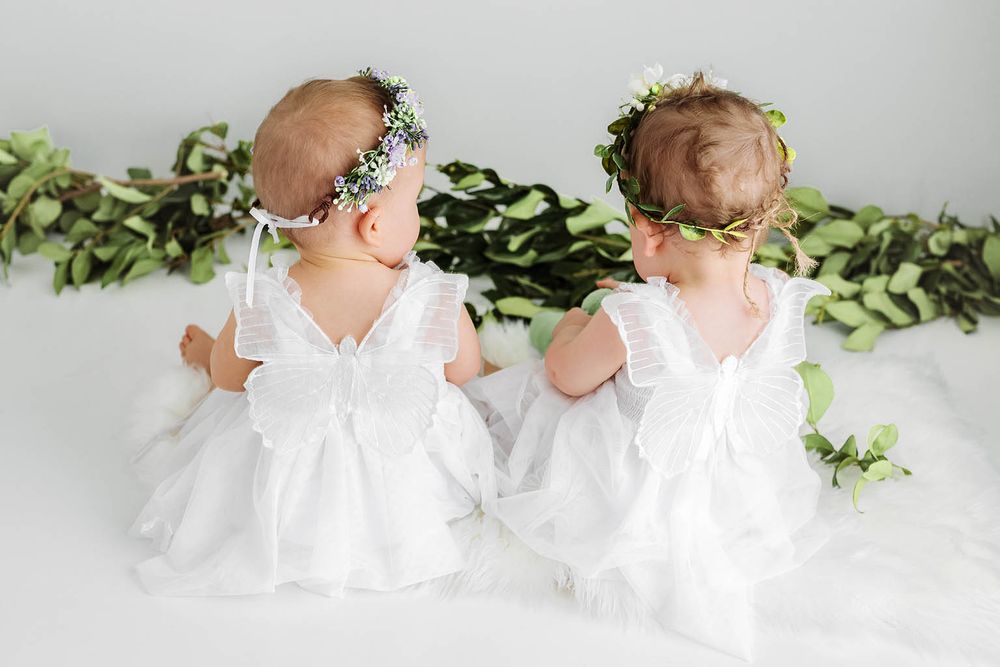 Two baby girls in white dresses and floral crowns sitting together, viewed from behind.