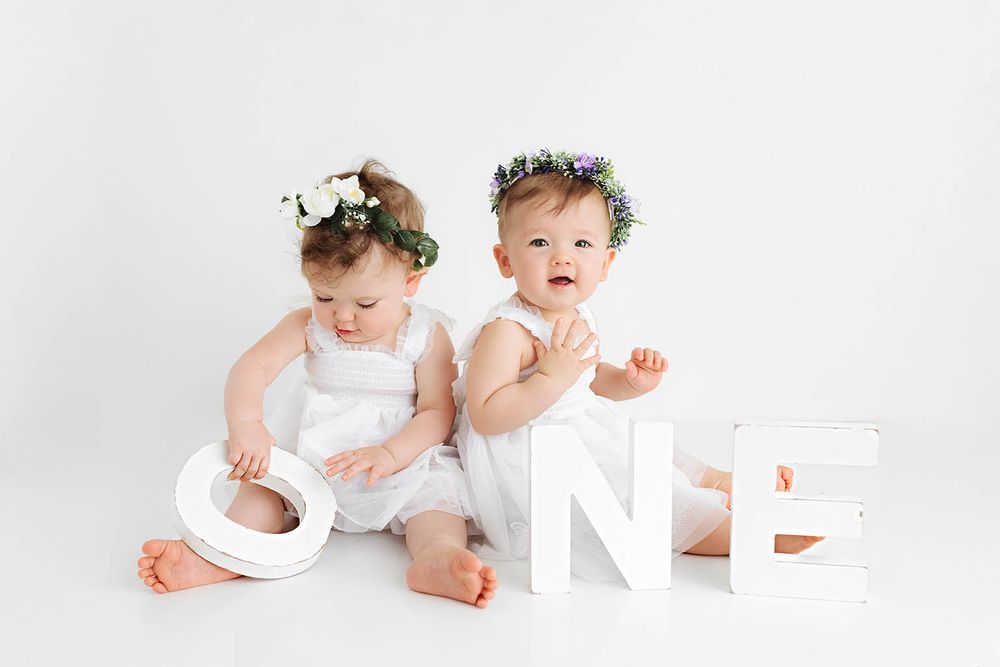 Two baby girls in white outfits and floral crowns pose with large ONE letters for their first birthday photo.