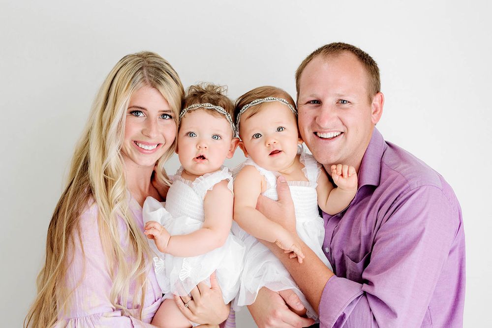 A smiling family with a mother, father, and twin baby girls in white dresses posing for a portrait photo.
