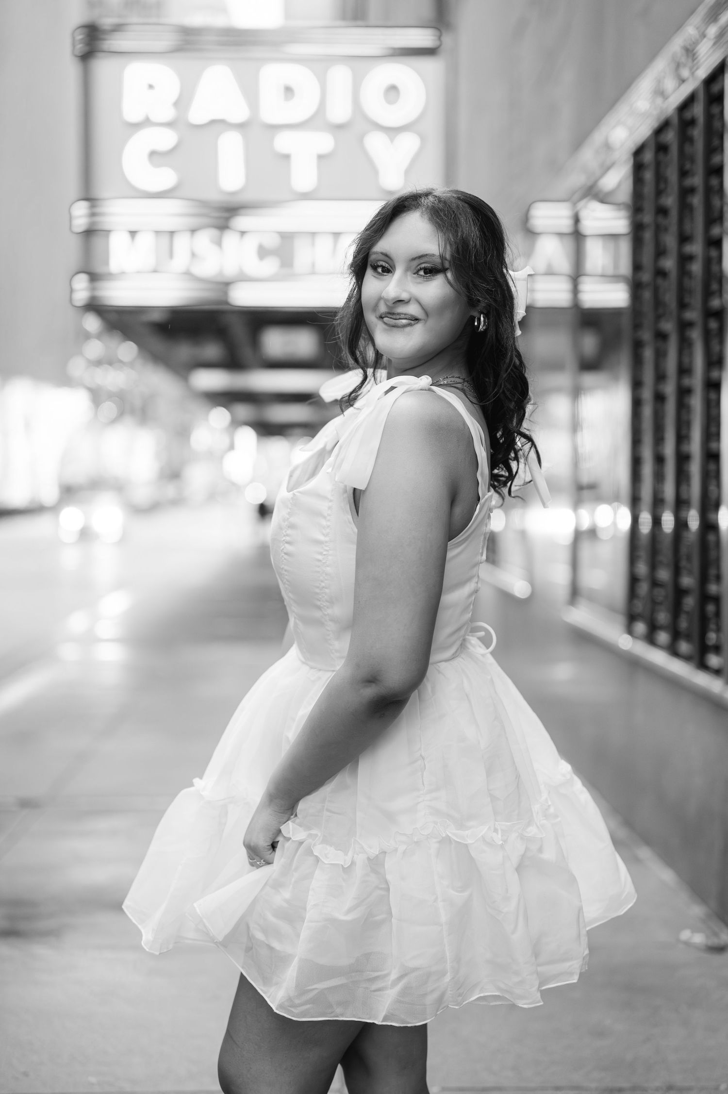 Young woman in white dress and heels poses on NYC sidewalk outside Radio City Music Hall during sweet sixteen portrait session.