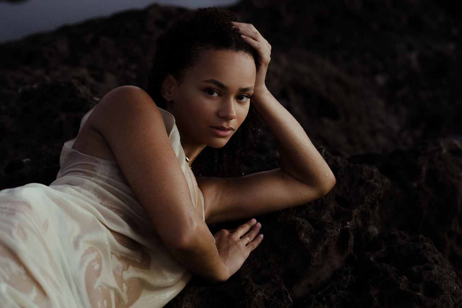 Young woman with curly hair leans on a railing outdoors, wearing a delicate gold necklace near the water.