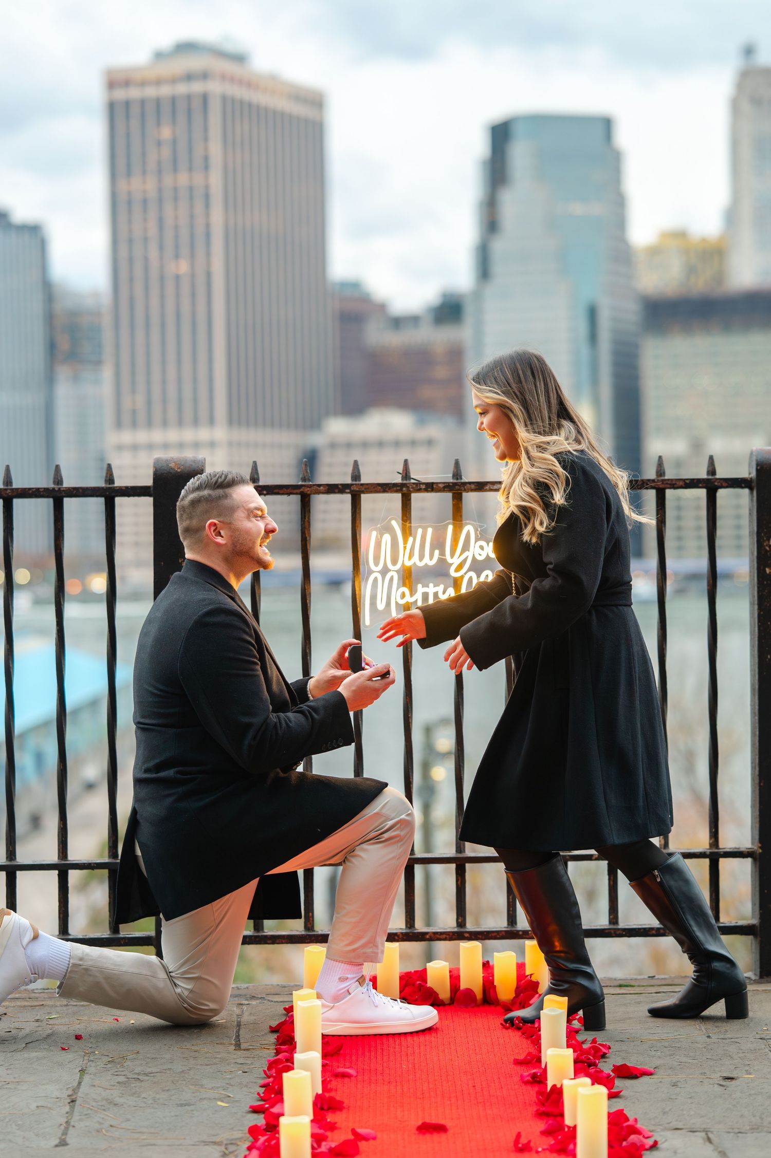 Man proposes to woman on one knee with candles and Will You Marry Me sign against NYC skyline backdrop.