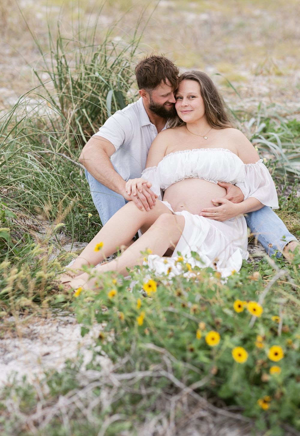 Man embraces expectant mother wearing cropped white outfit while both sitting in coastal grass. Hampton York maternity photographer