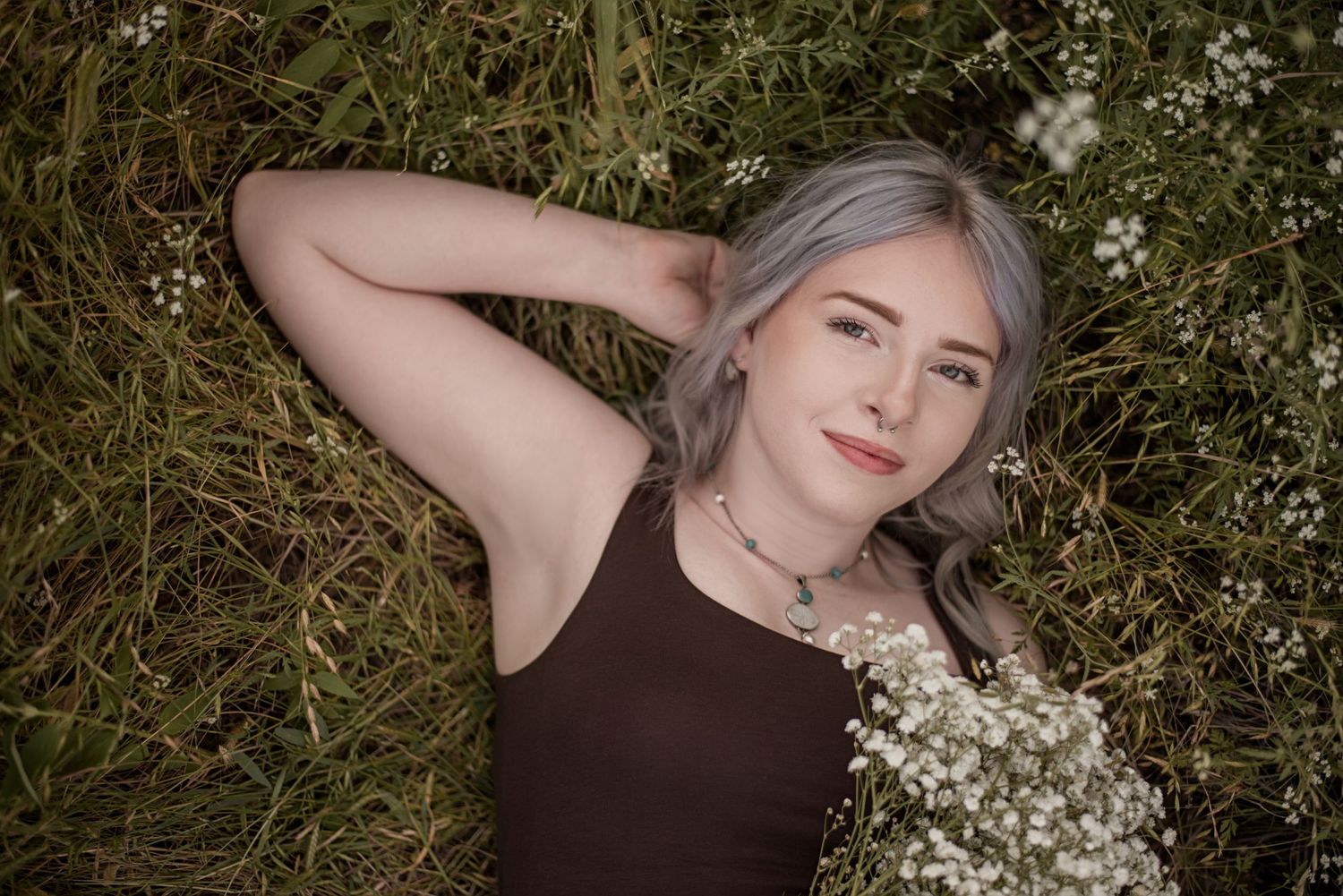 Young woman with silver hair lying in green grass holding white wildflowers, wearing a brown top and delicate necklace.