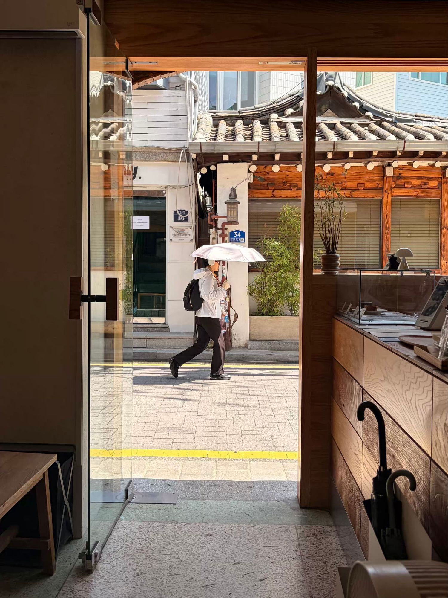 Person walking with white umbrella past traditional Korean architecture, viewed through glass doorway.
