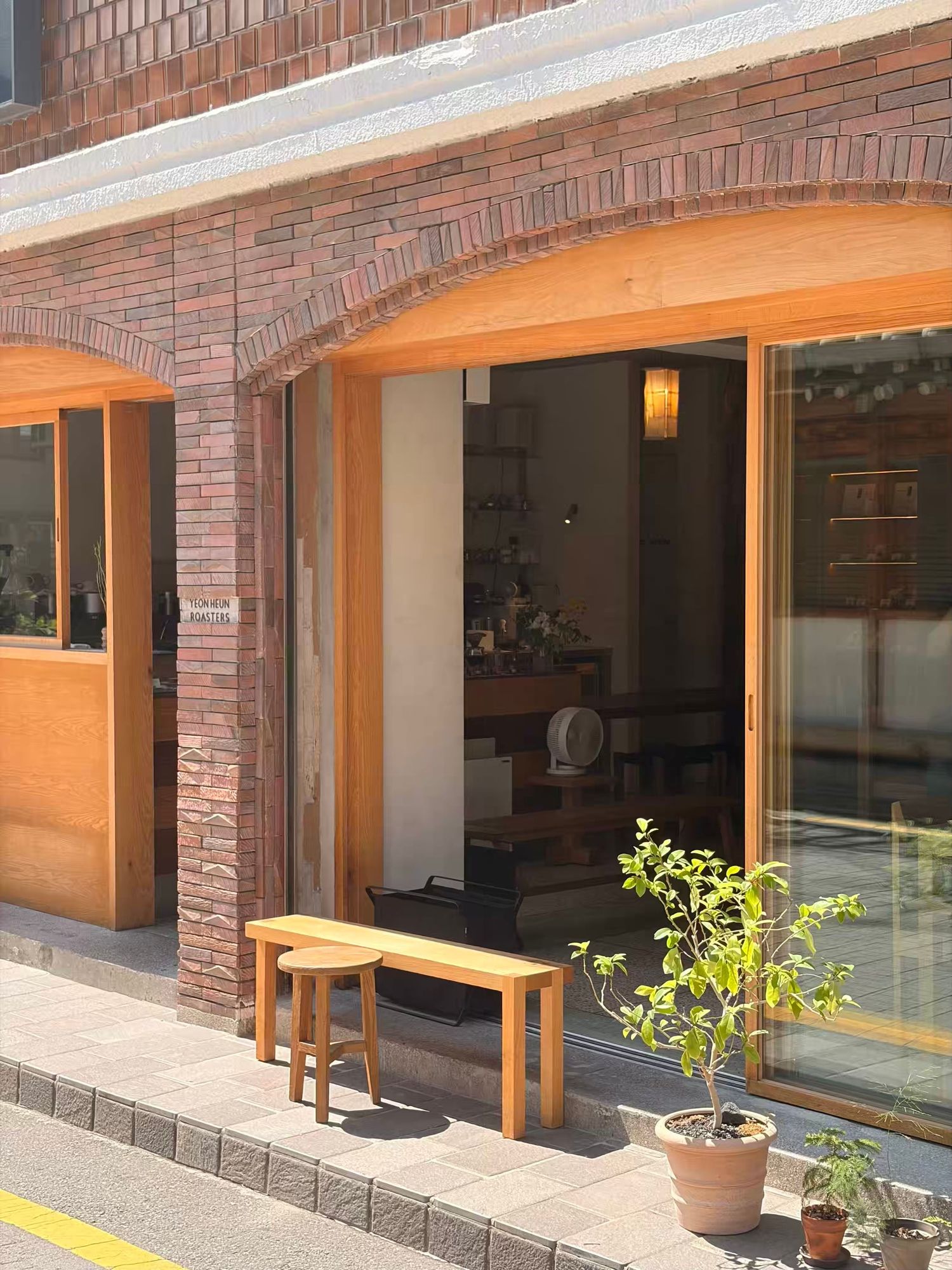 Cozy cafe exterior with brick facade, wooden benches, and potted plant by open glass doors.