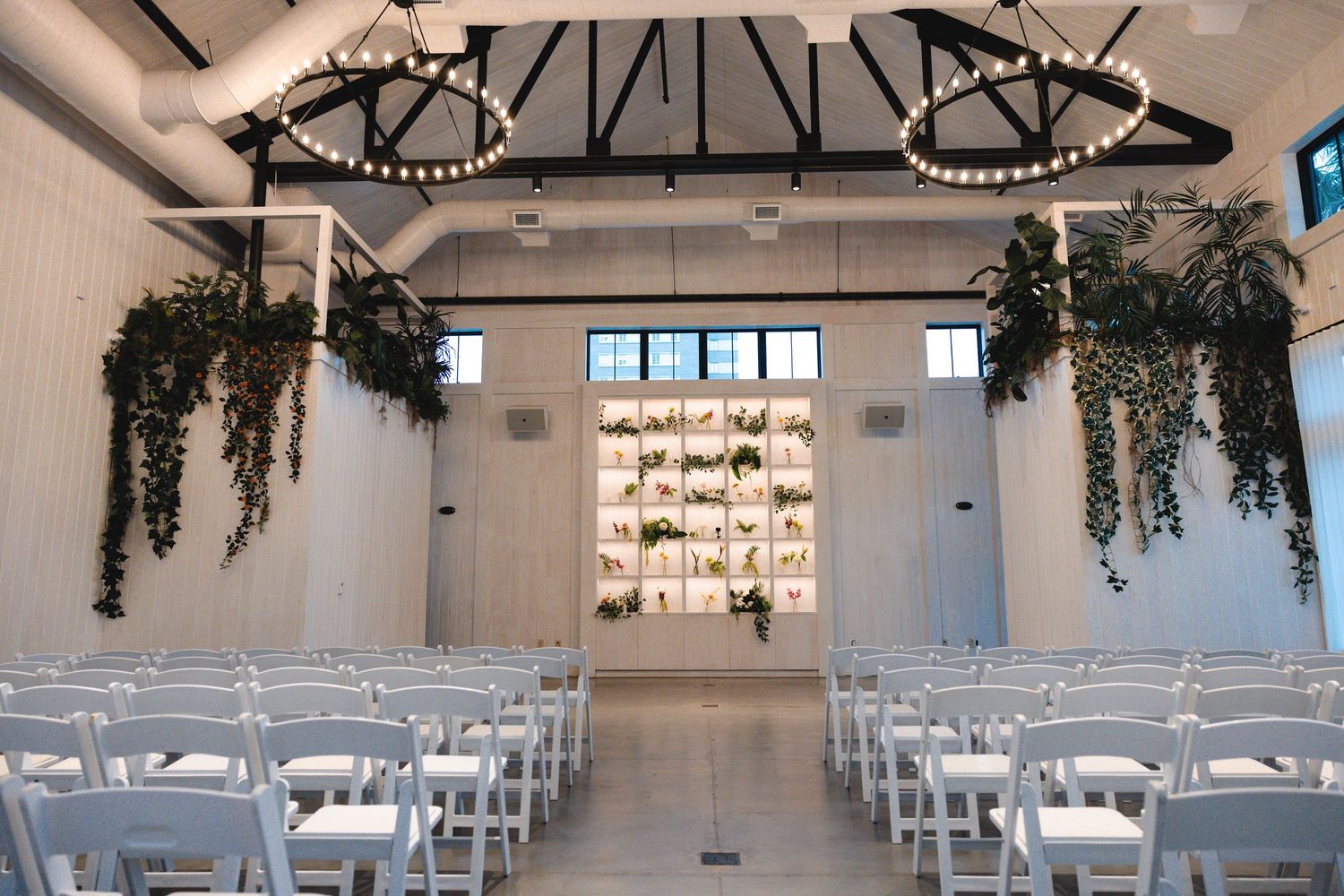 Elegant wedding ceremony venue with white chairs, hanging greenery, chandelier, and a botanical backdrop wall.