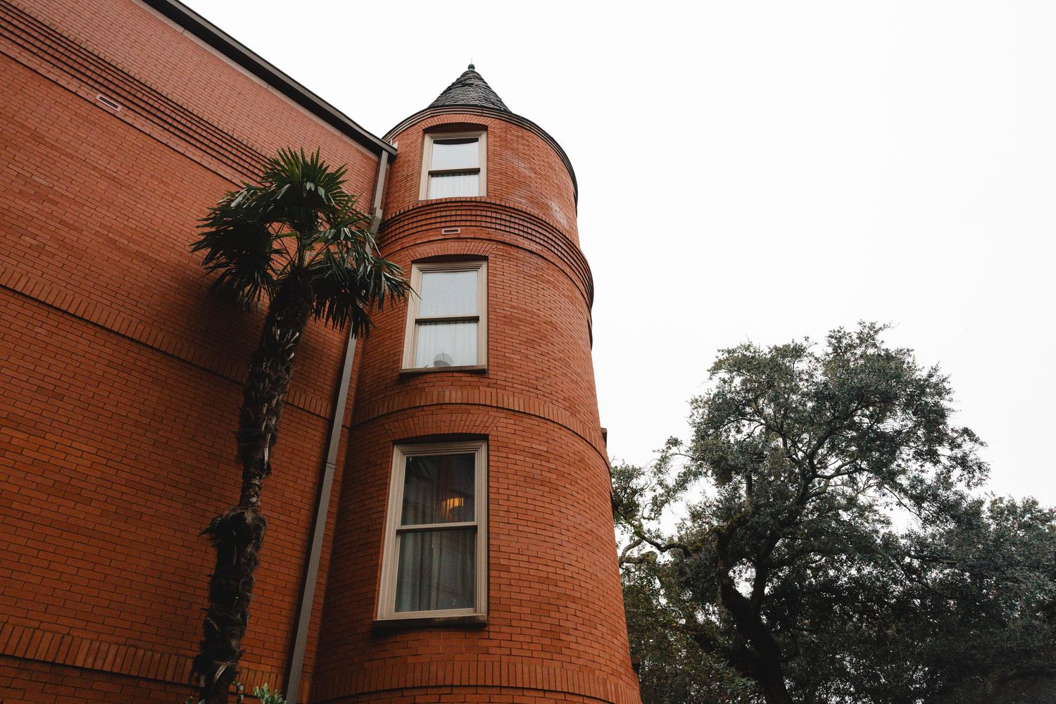 Red brick Victorian building with rounded turret, palm tree, and large oak tree against overcast sky.