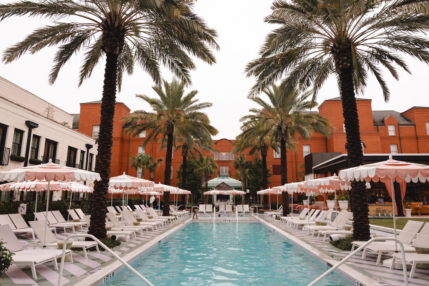 Luxury hotel pool surrounded by tall palm trees, pink umbrellas, and white lounge chairs with a red building backdrop.