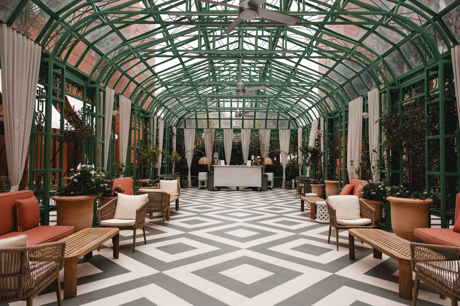 Elegant greenhouse conservatory with arched glass ceiling, wicker chairs, coral cushions, and geometric tile floors.