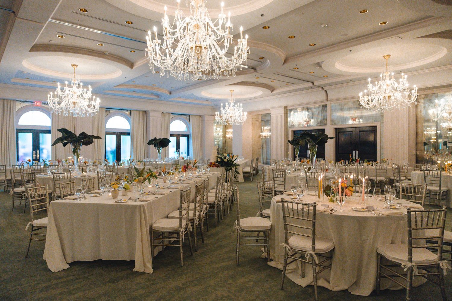 Elegant ballroom wedding reception with crystal chandeliers, long white tables, and gold chiavari chairs in a grand venue.