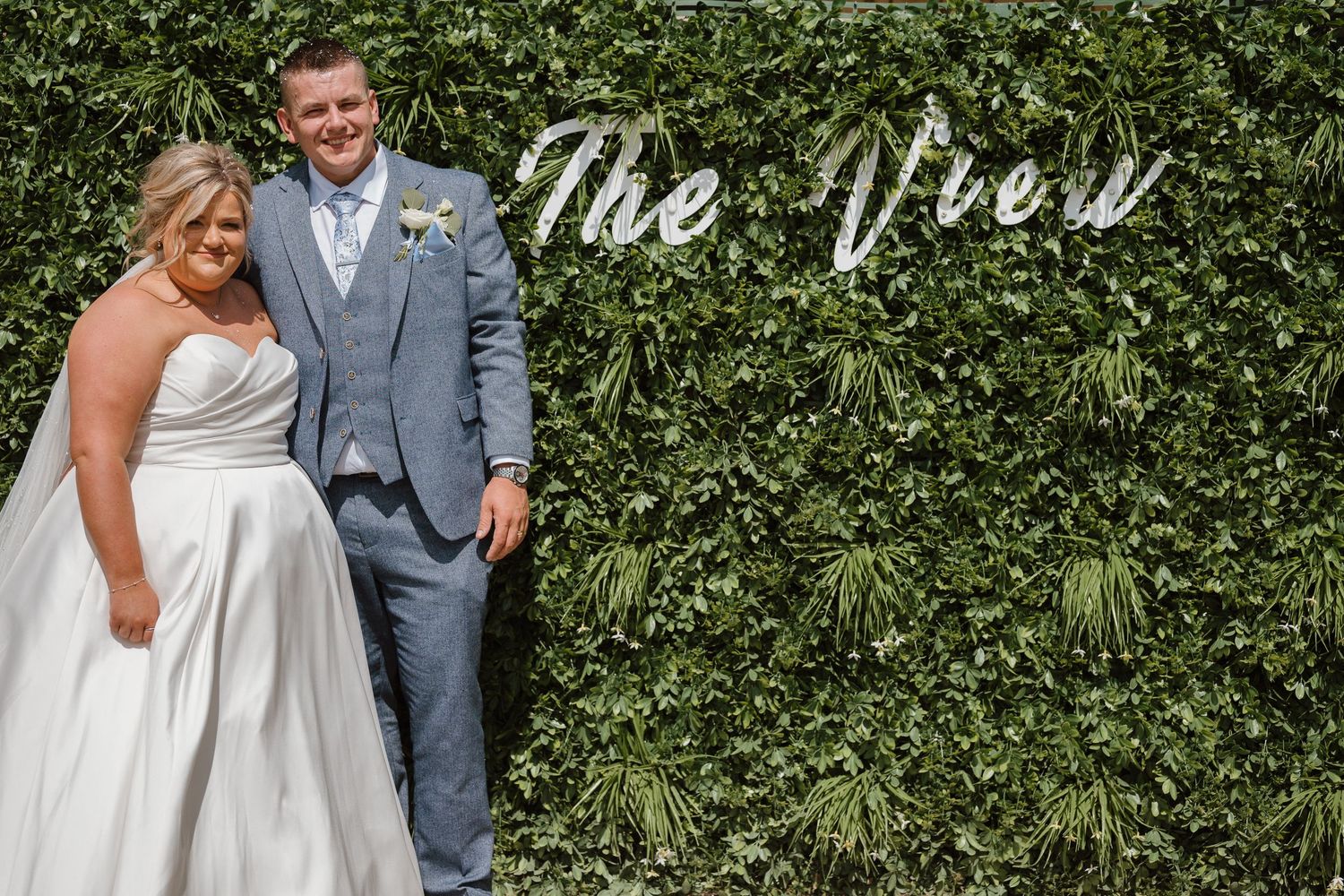 Bride and groom smiling in front of a lush green ivy wall with The View sign at their wedding venue.