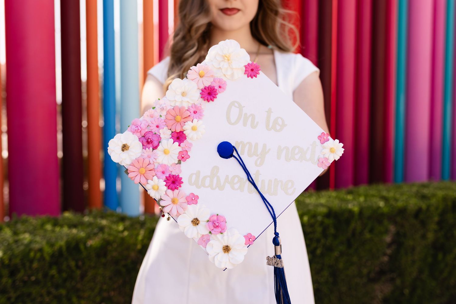 Graduate in white dress holds floral-decorated cap reading On to my next adventure with colorful striped backdrop.