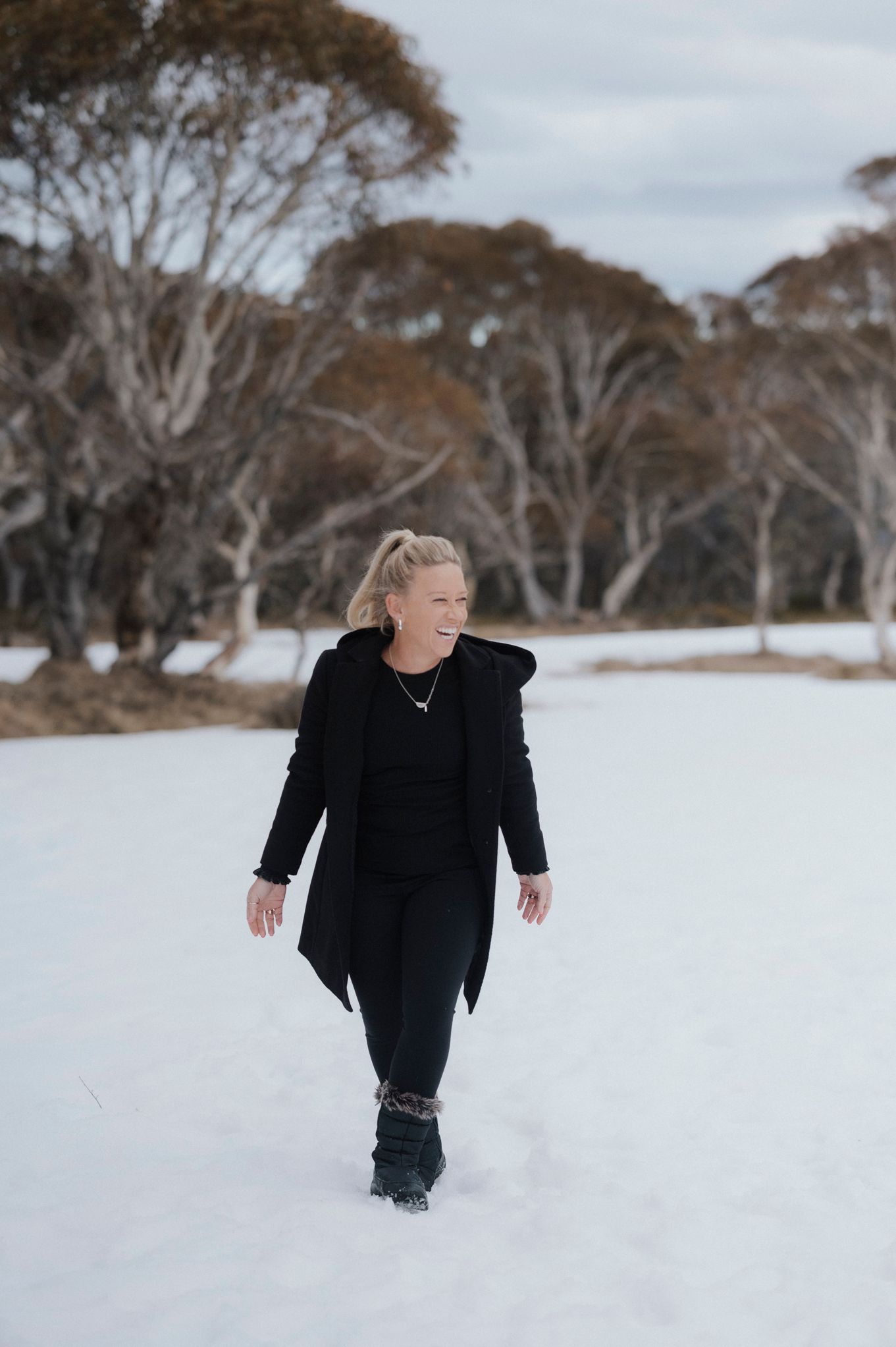 Woman in black outfit walking through snow with bare winter trees in background, smiling outdoors.