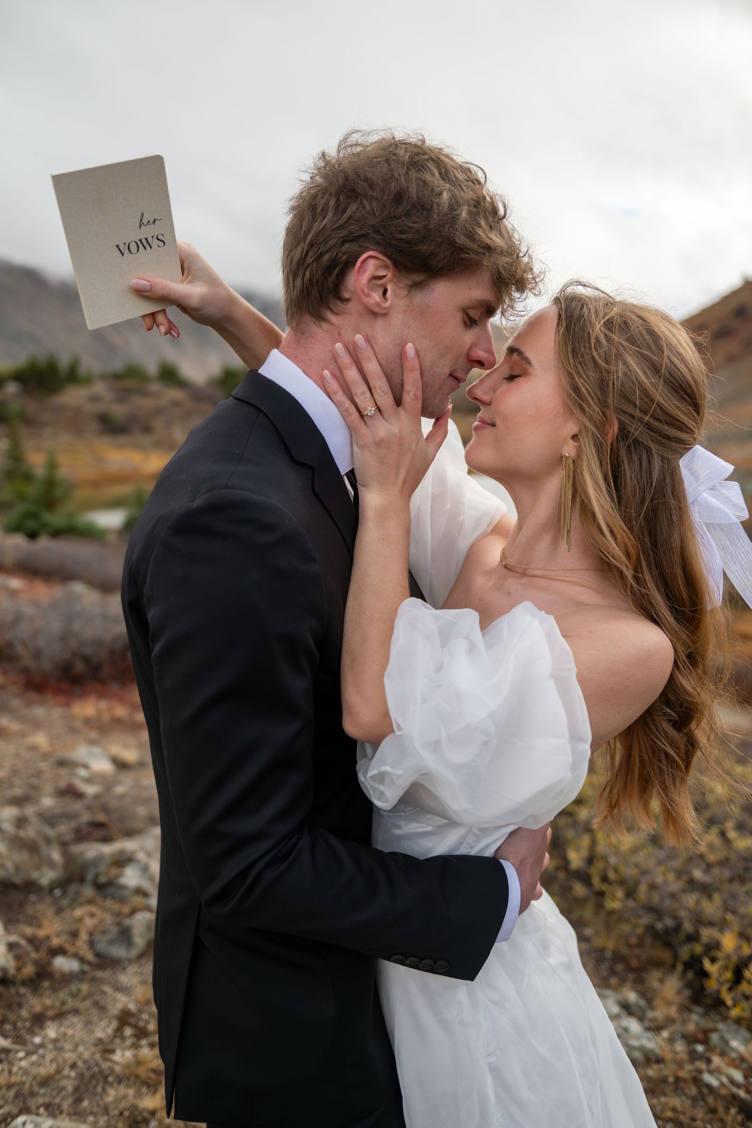 Bride and groom share a romantic moment outdoors, she holds vow booklet, wearing white gown, he in black suit, mountains behind.