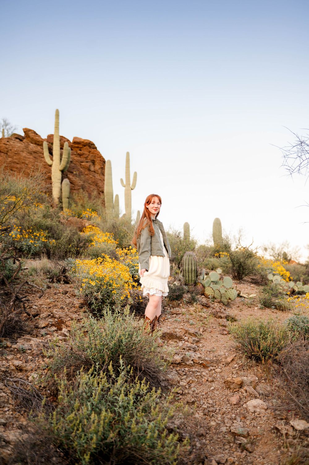 Woman in white skirt and green jacket walking through Arizona desert with saguaro cacti and yellow wildflowers.
