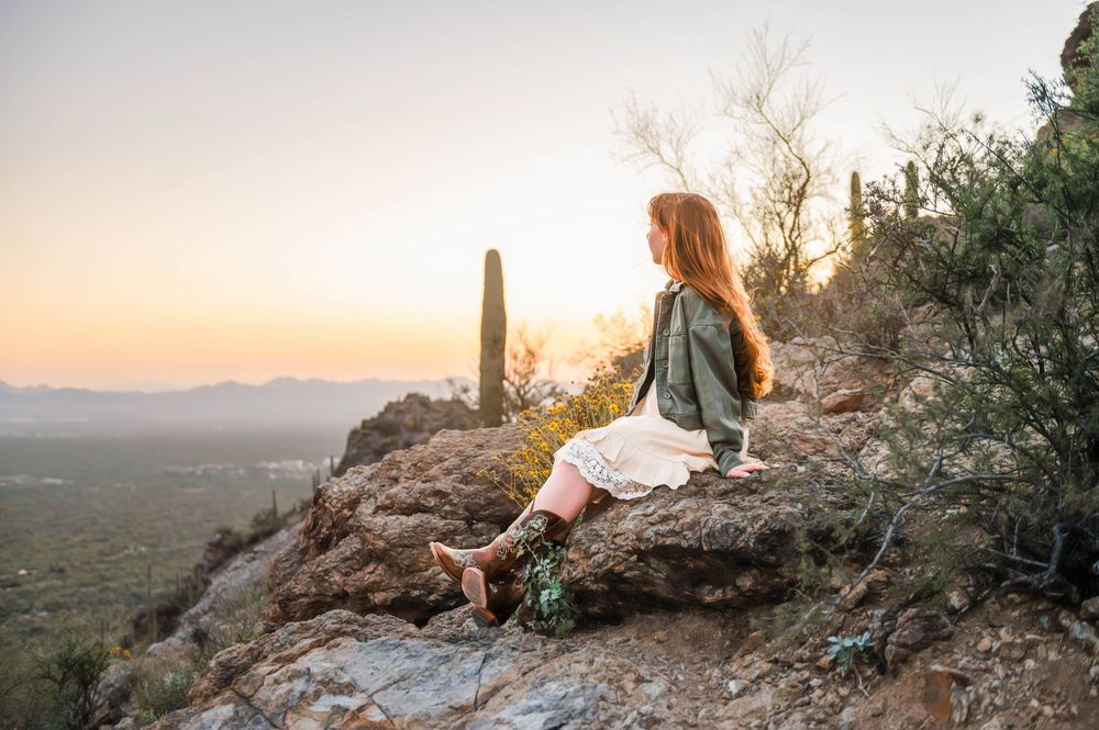 Young woman in white skirt sitting on rocky hilltop overlooking desert valley at golden sunset.