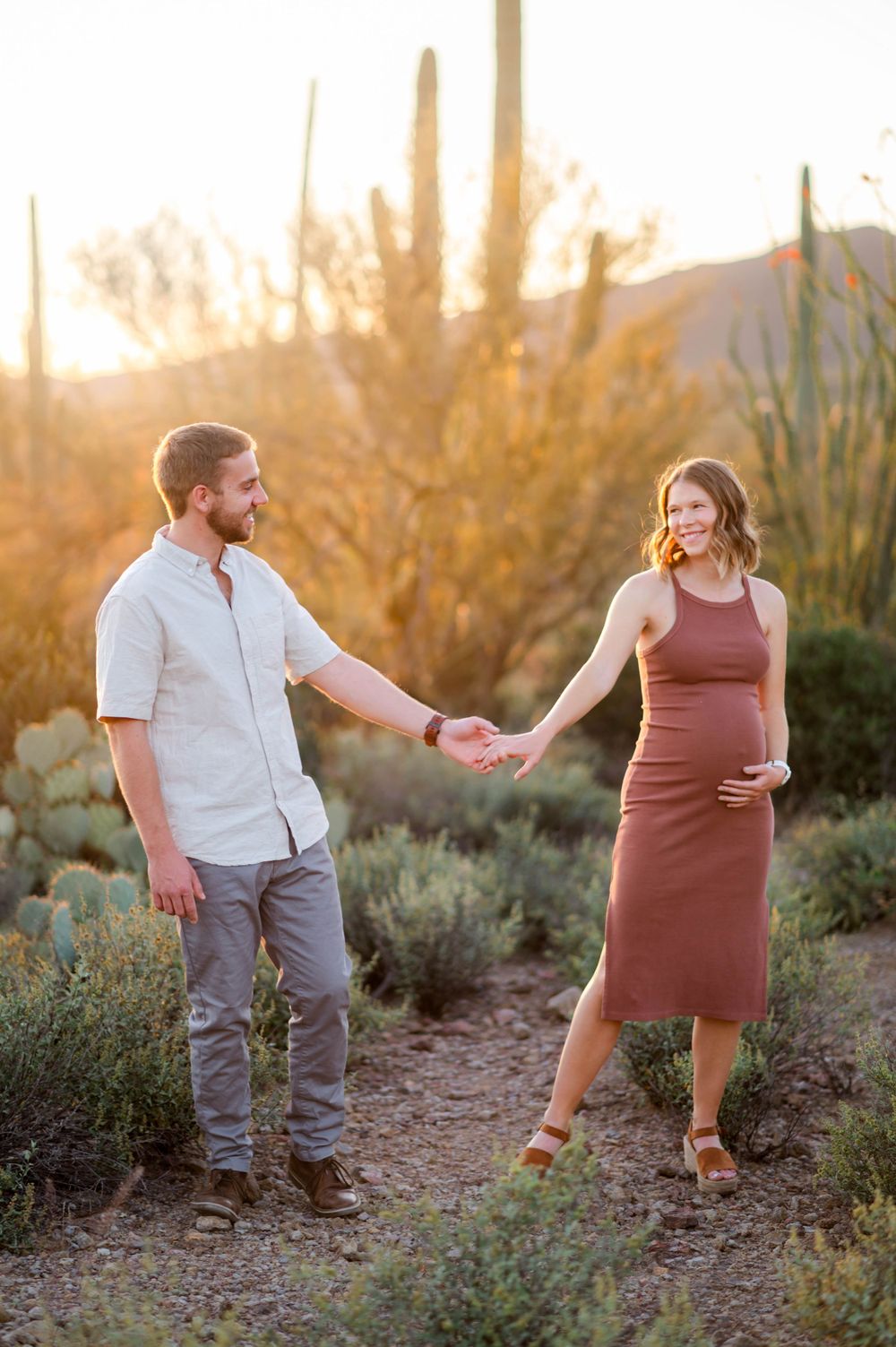 Expecting couple holds hands in a glowing Arizona desert sunset surrounded by saguaro cacti.