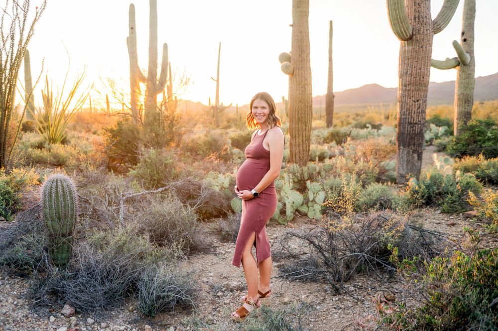 Pregnant woman cradles her bump in a warm golden Arizona desert sunset with saguaro cacti.