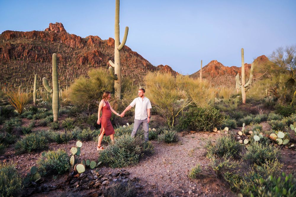 Couple holds hands while walking through a scenic Arizona desert with saguaro cacti and red mountains.