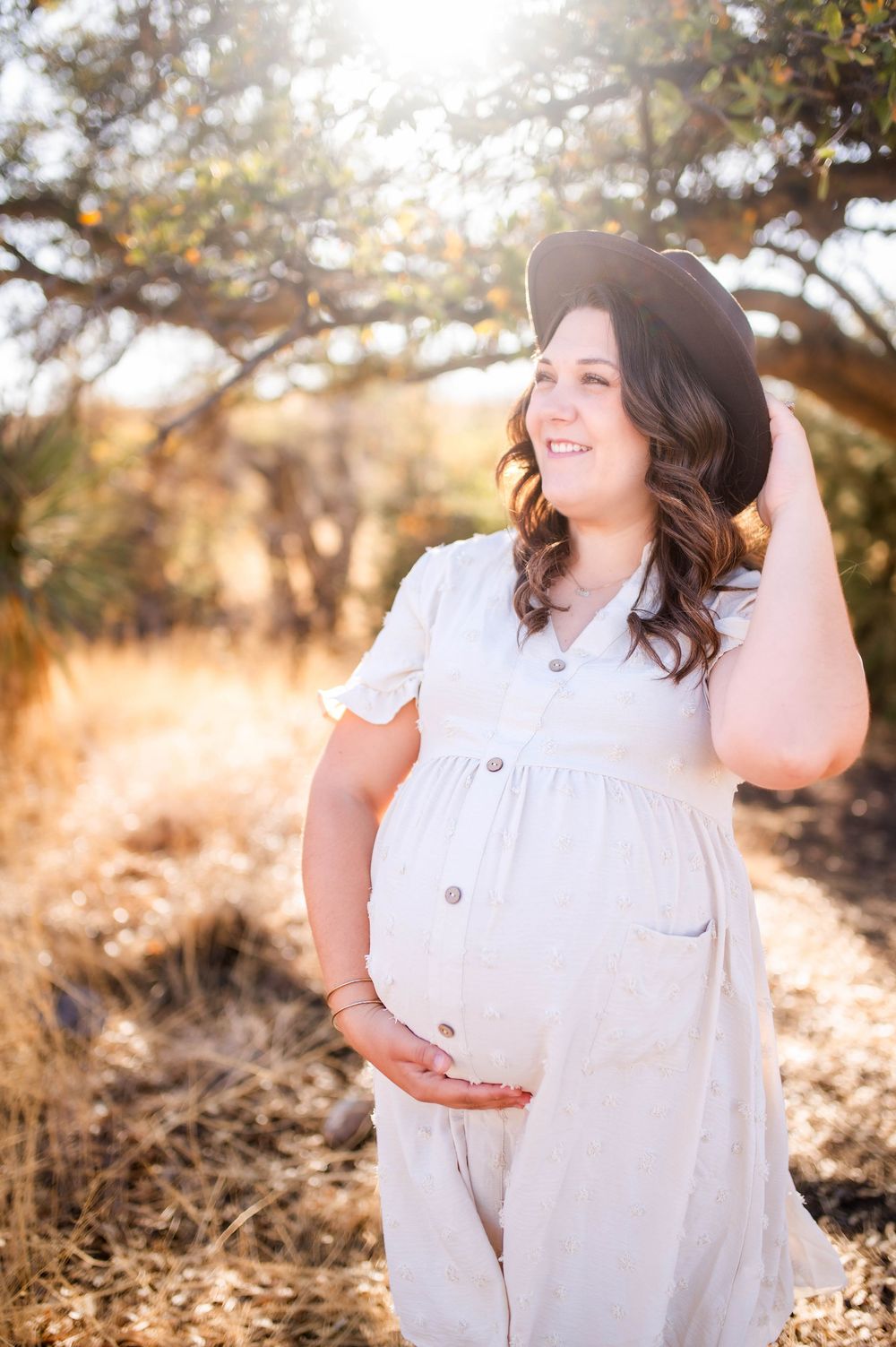 Smiling pregnant woman in white dress and black hat cradling baby bump in sunlit field.