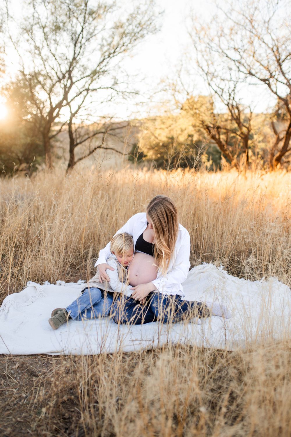 Pregnant mother sitting on white blanket in golden field while toddler kisses her bare baby bump.