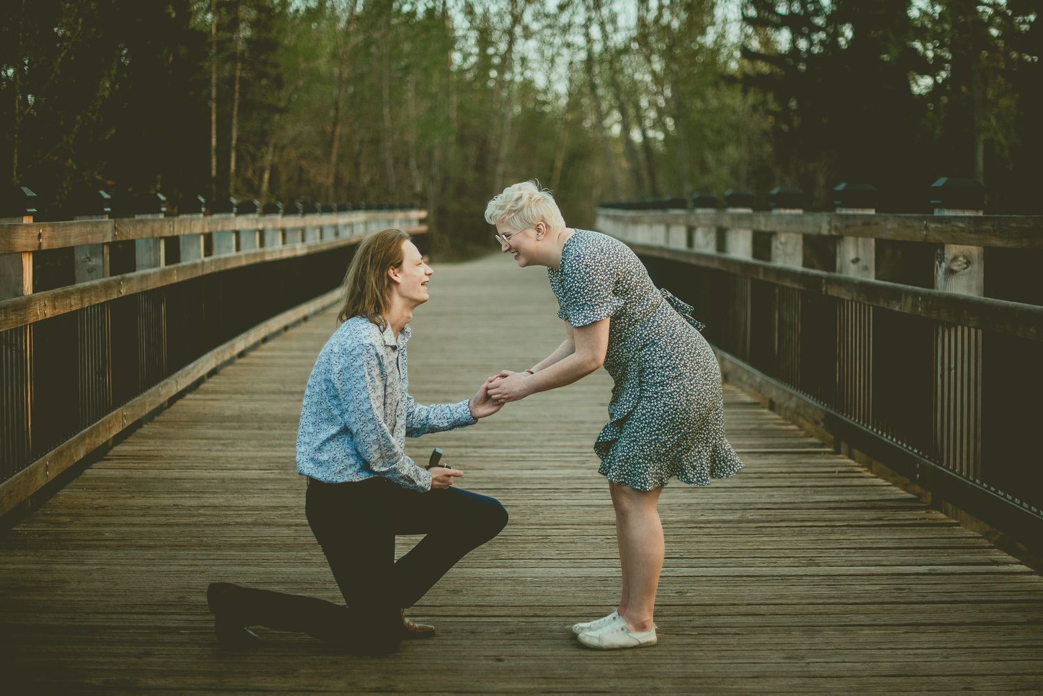 A person kneeling on a wooden bridge proposing to another person outdoors surrounded by trees.