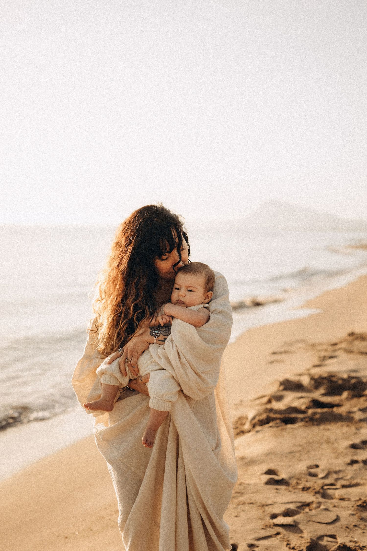 Mother with curly hair tenderly holds baby on sunny beach, wrapped in cream blanket with ocean waves behind Javea photographer