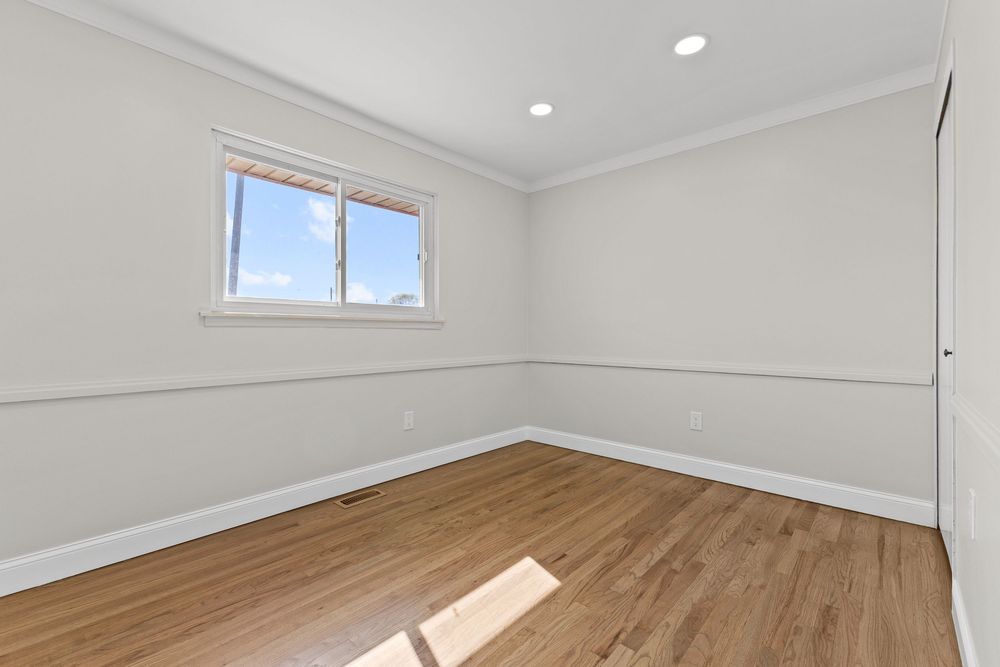 Bright empty bedroom with hardwood floors, crown molding, recessed lighting, and large sunny window.