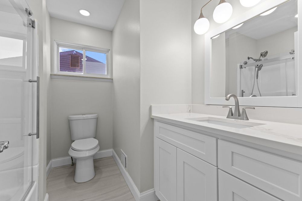 Clean white bathroom with single vanity, large mirror, toilet, and walk-in shower with window.