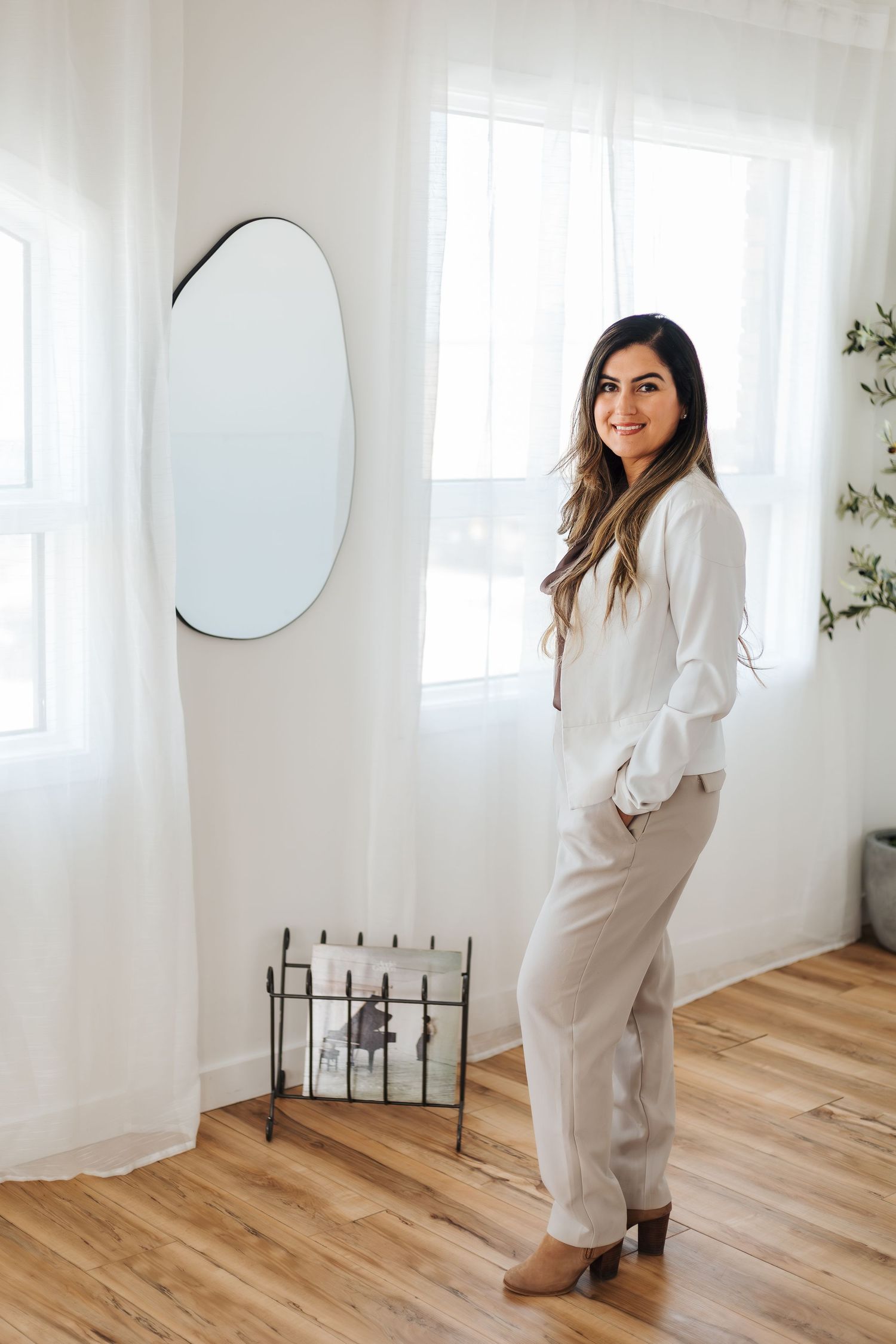 A smiling woman in white blouse and beige trousers poses in a bright minimalist room with oval mirror and wood floors. Luxury branding