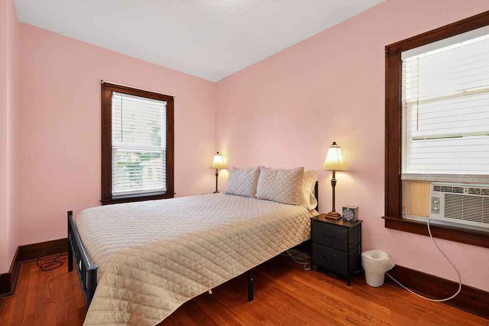Pink bedroom with hardwood floors, black metal bed frame, two windows, and wall AC unit.