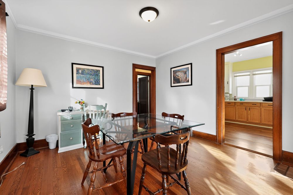 Elegant dining room with hardwood floors, glass table, wooden chairs, crown molding, and view into sunlit kitchen.