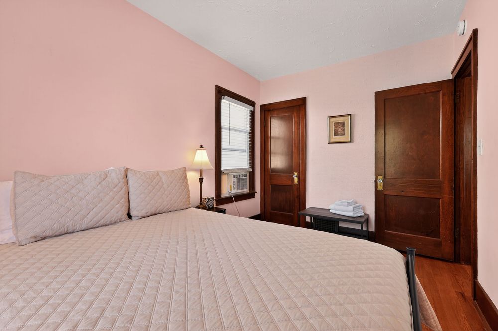 Cozy pink bedroom with queen bed, hardwood floors, wooden door, and window with natural light.