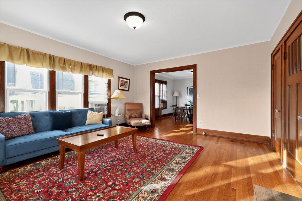 Bright living room with hardwood floors, blue sofa, red area rug, crown molding, and view into dining room.