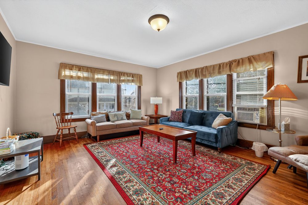 Bright living room with hardwood floors, blue sofa, red area rug, and multiple windows with curtains.