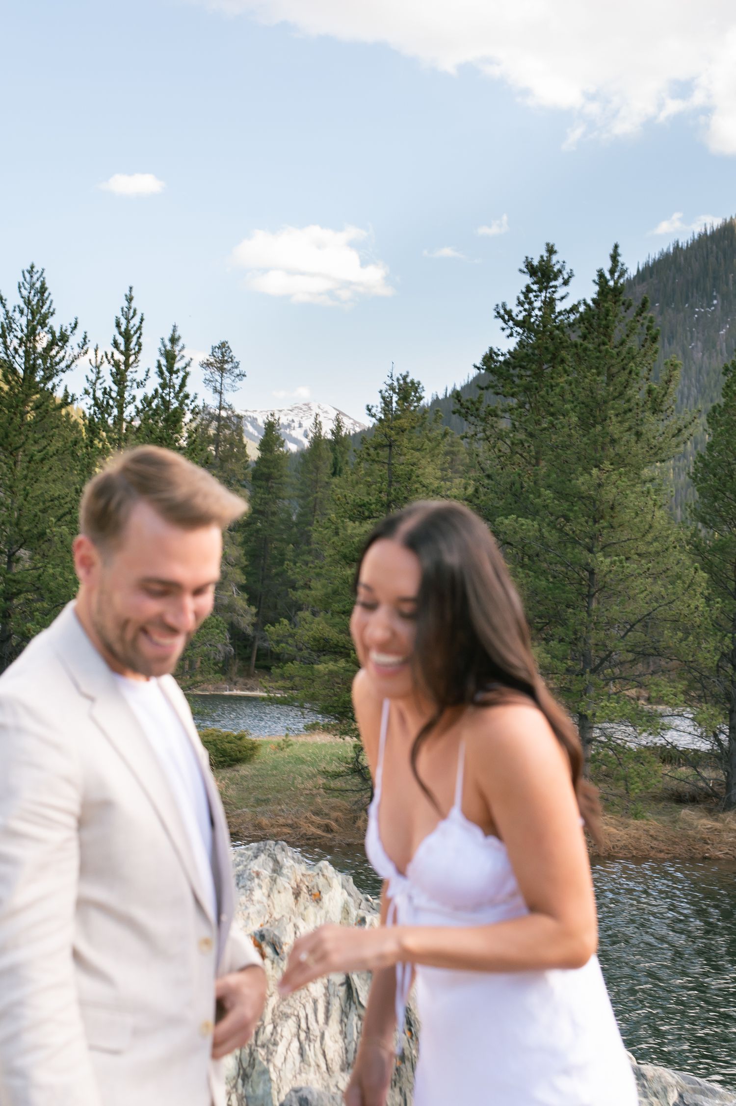 Couple in white attire laughing near a mountain lake surrounded by pine trees during an outdoor engagement session.