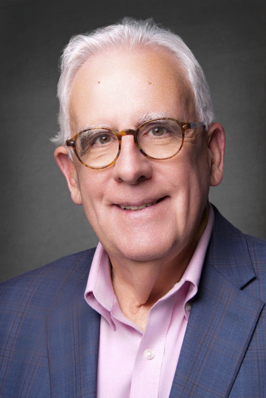 Professional headshot of a smiling older man with white hair, tortoiseshell glasses, a blue blazer and pink shirt.