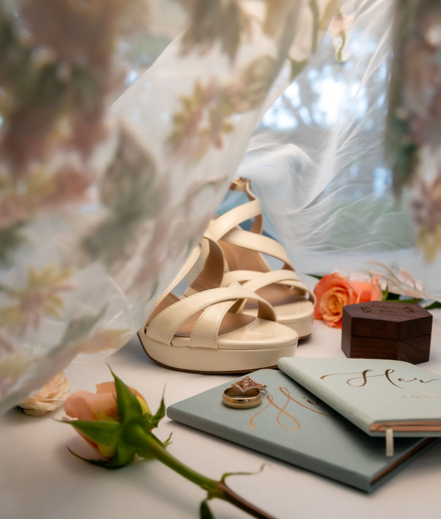 Elegant bridal flat sandals with cross straps beside roses, a ring, and a sage green journal on a white surface.