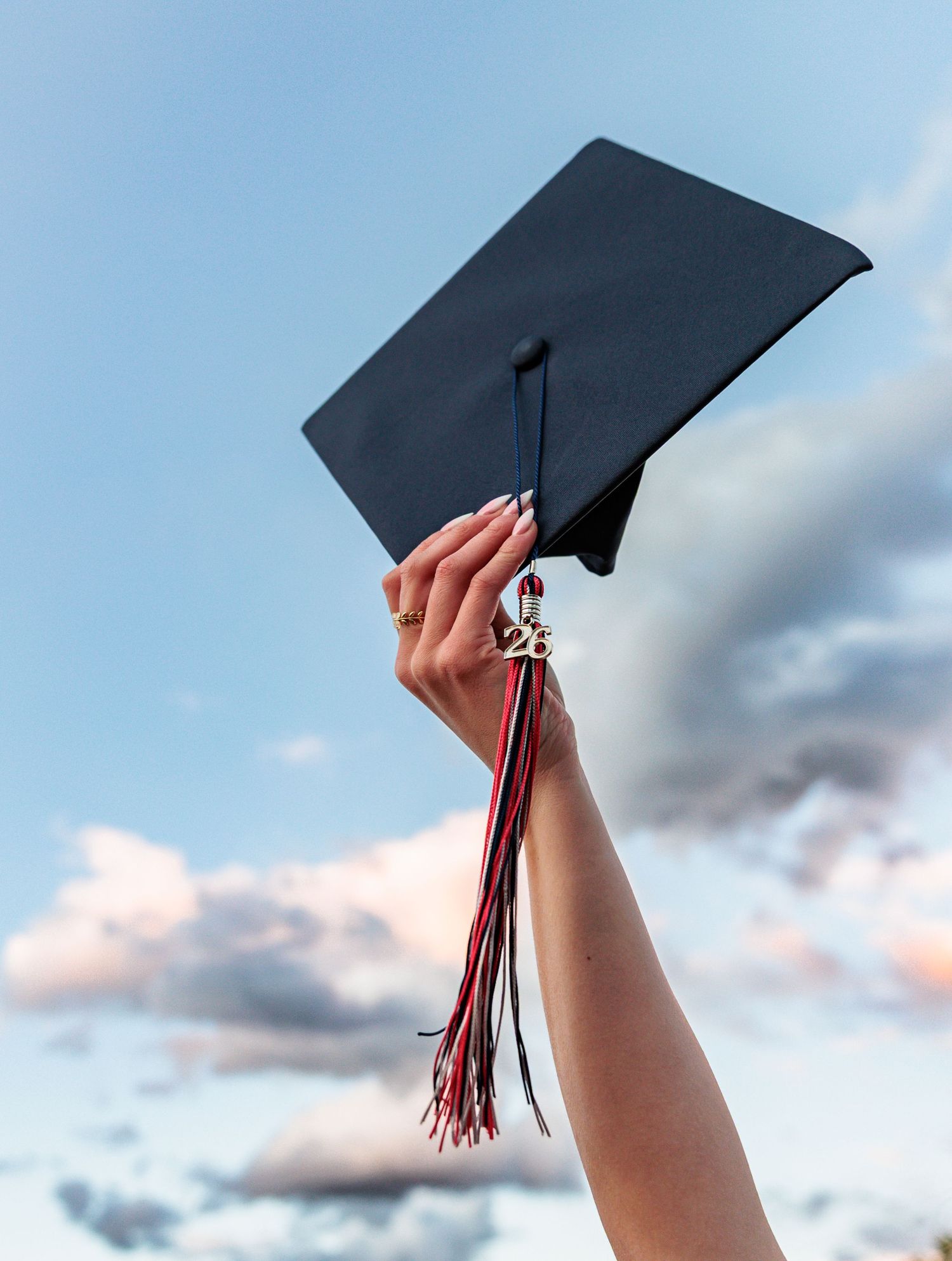A hand raises a black graduation mortarboard cap with red and black tassel against a blue cloudy sky.