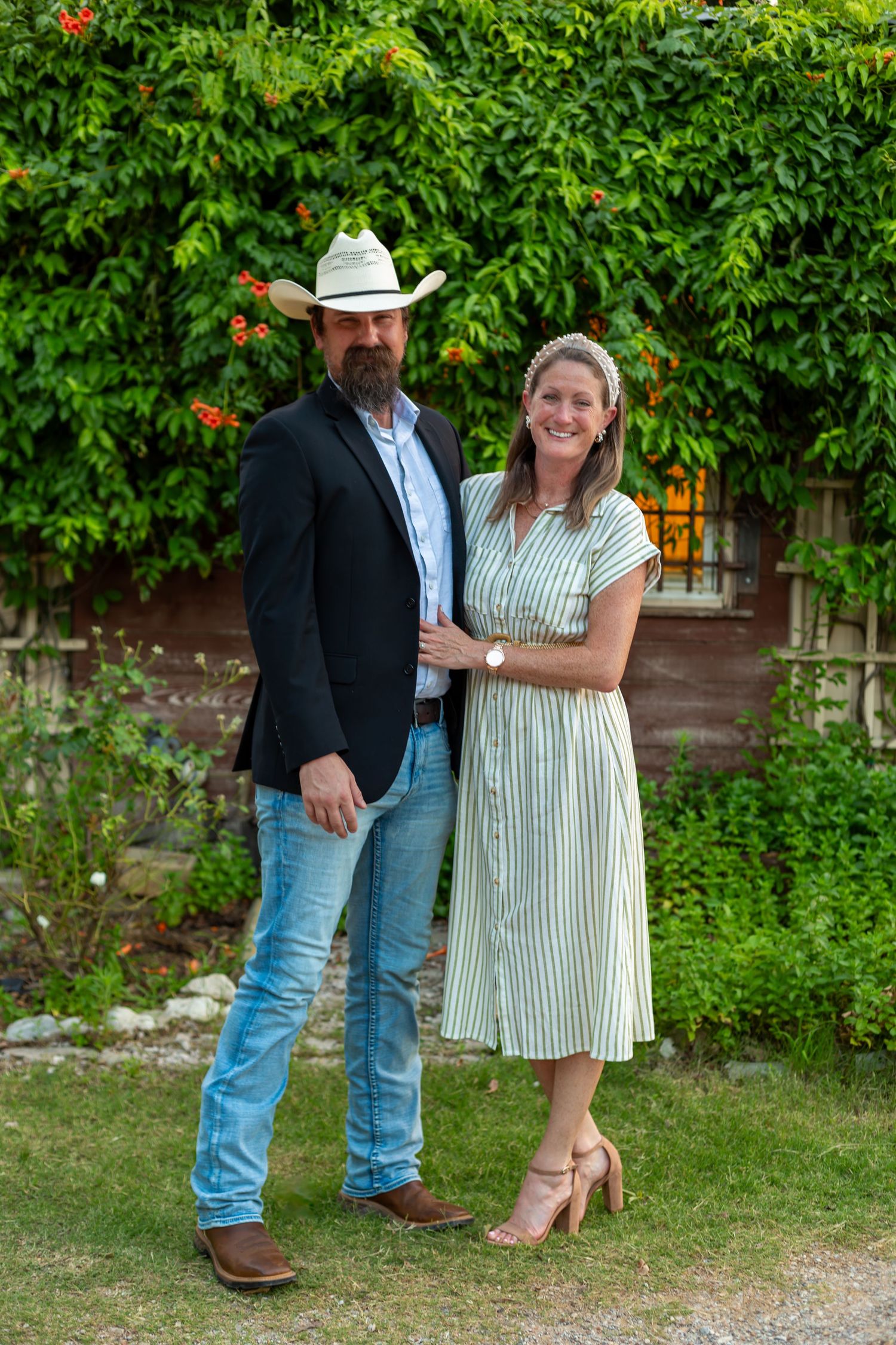 A couple poses outdoors in front of lush greenery, man in cowboy hat and blazer, woman in striped dress.